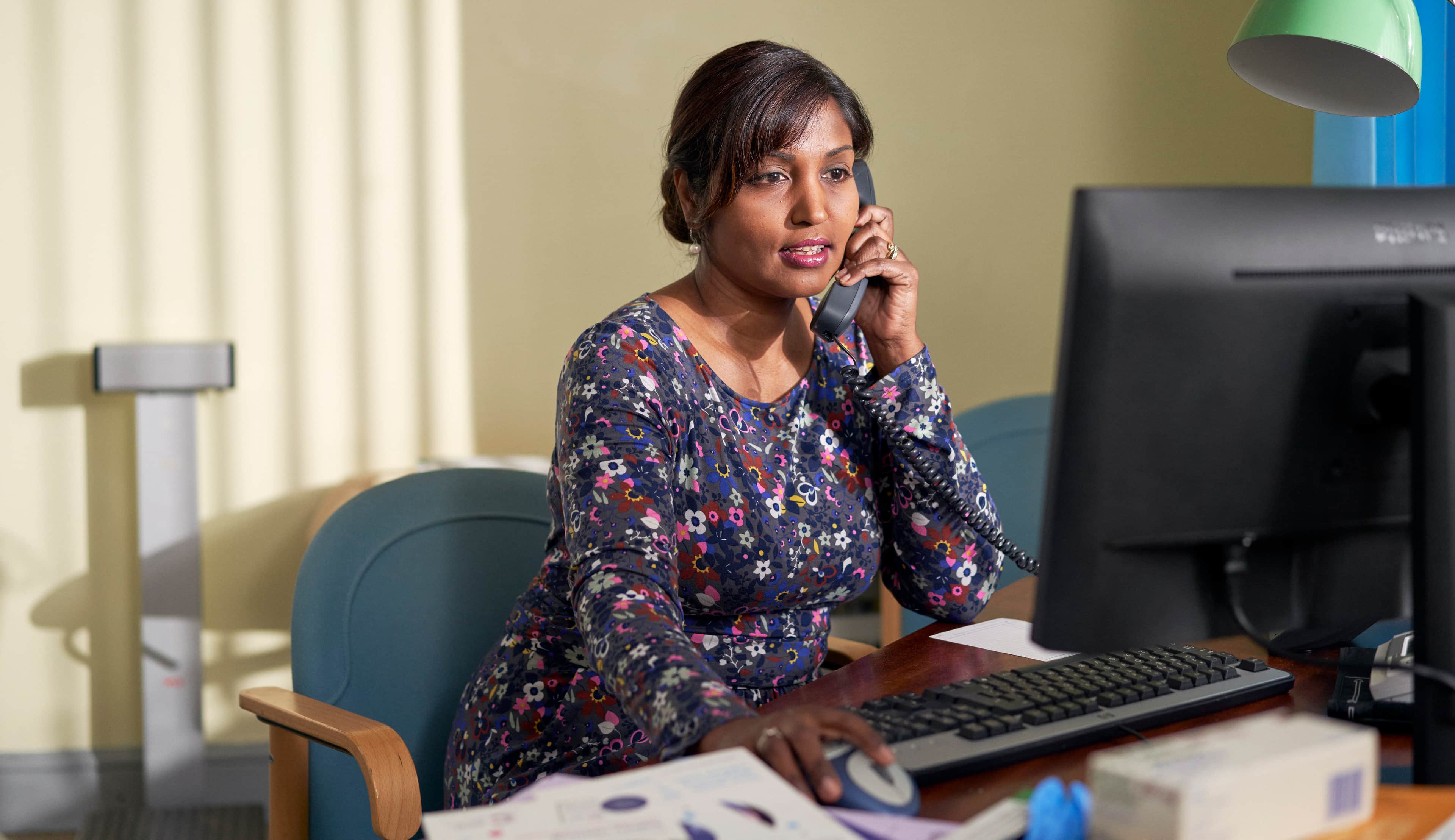 A GP is in her office, speaking on the phone and looking at her computer.