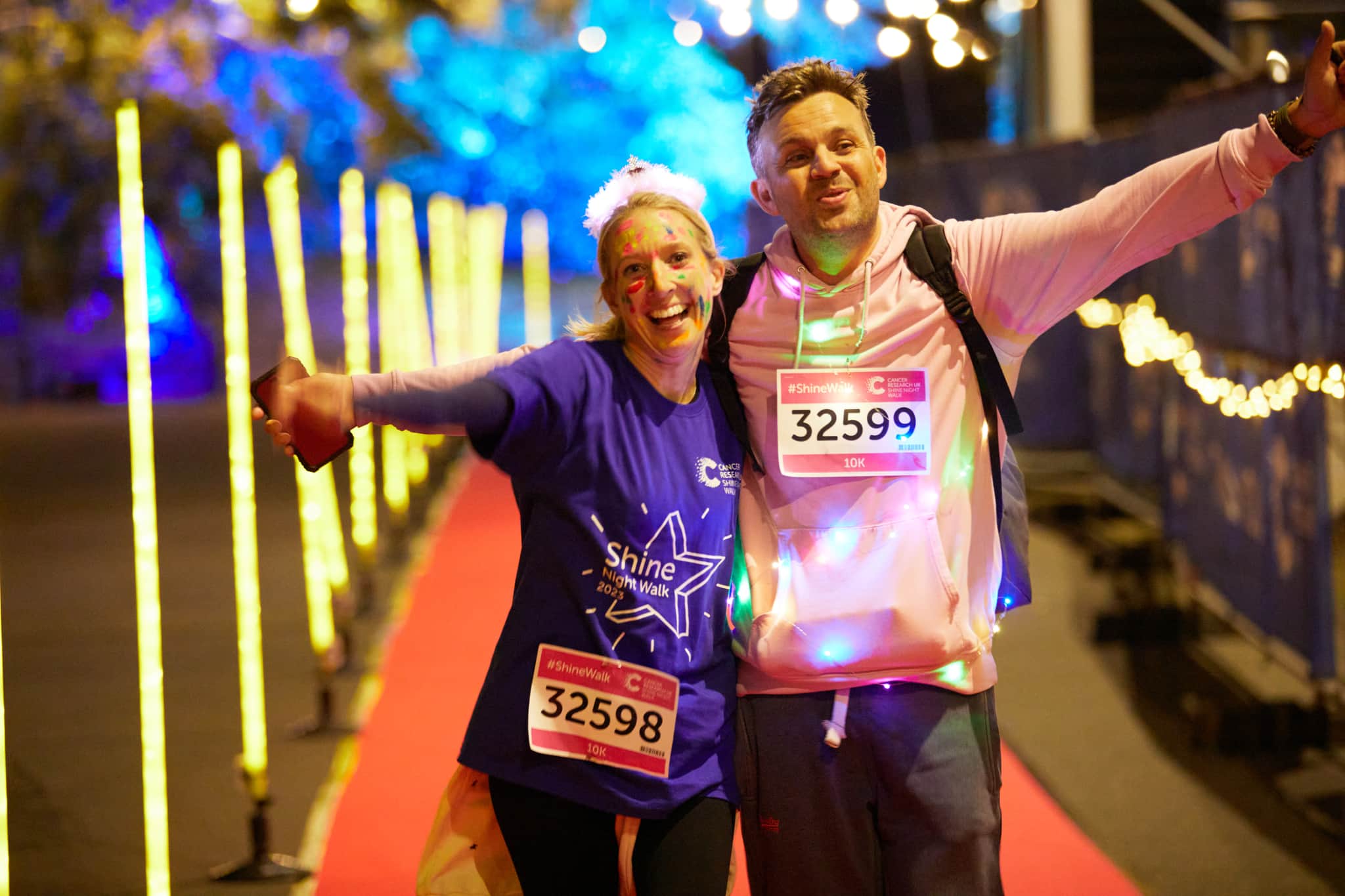 Two shine night walk participants wearing face paint and a fluffy headband walking down a red carpet lined with lights.