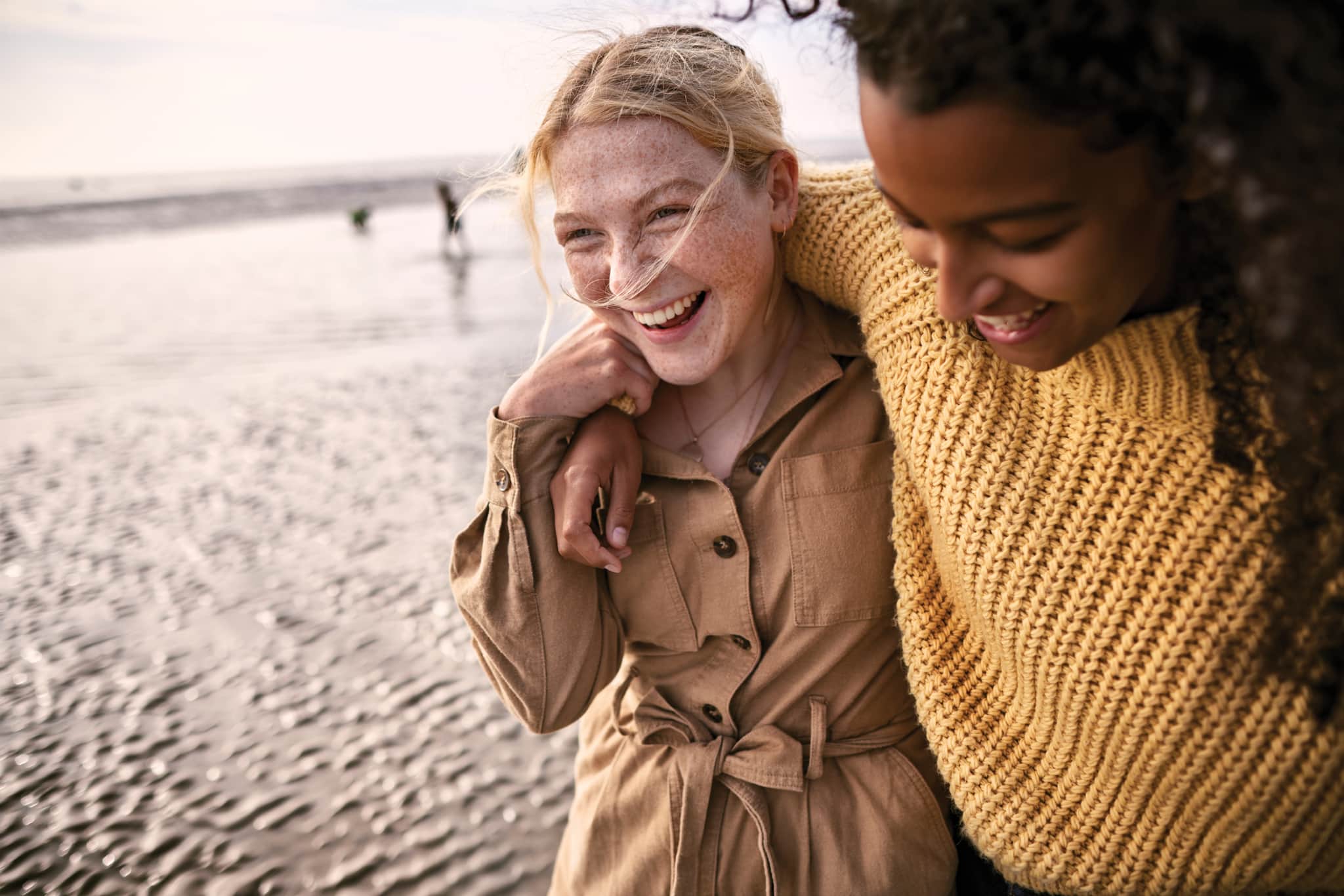 A photo of two women walking on the beach laughing and hugging.