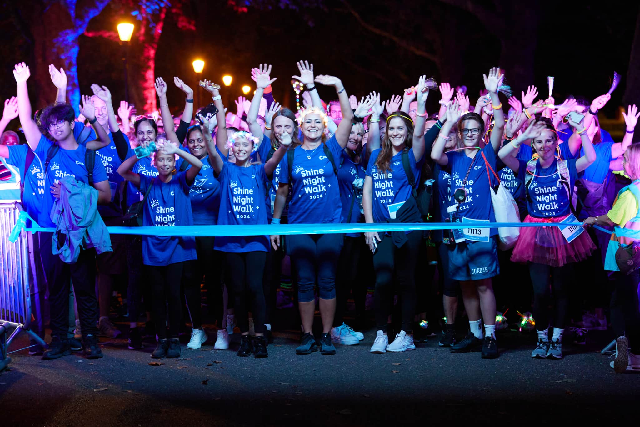 A group of smiling shine night walk participants celebrating with their hands up in the air and a blue finish line ribbon.