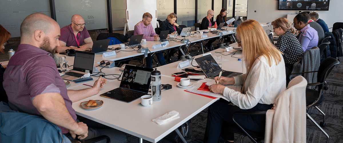 Eleven people sat around a rectangular desk with laptops at panel meeting.