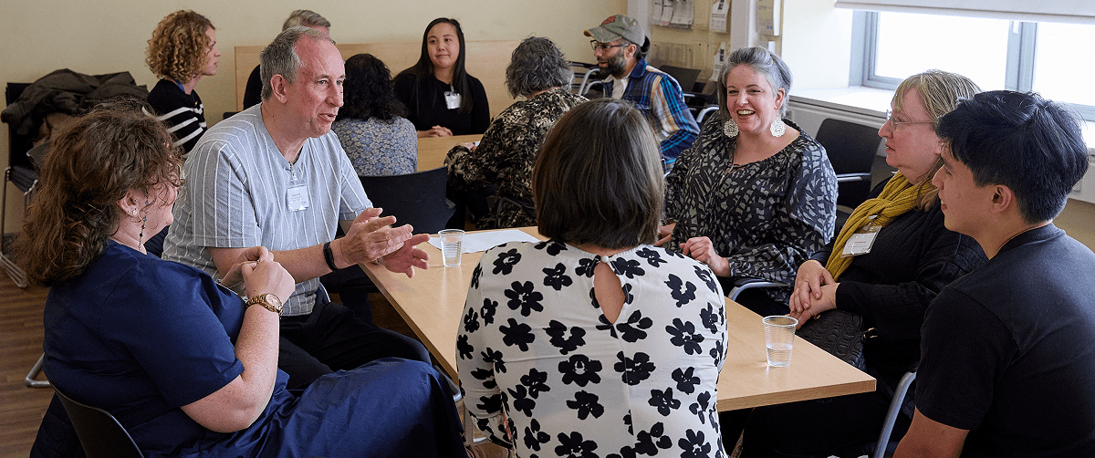 A group of people in a room. They are sat at tables having a discussion.