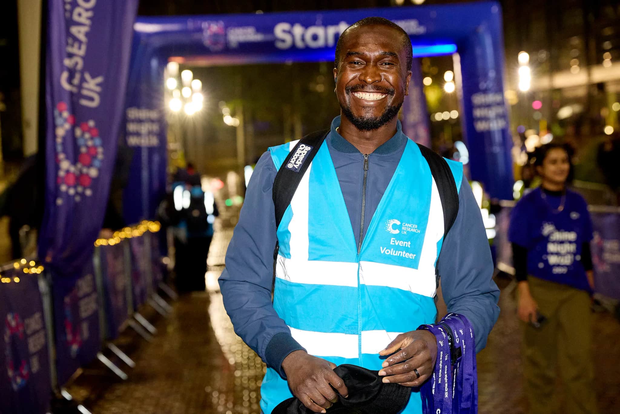 Image of a male volunteer, standing in front of the finish line at Shine Night Walk and smiling.
