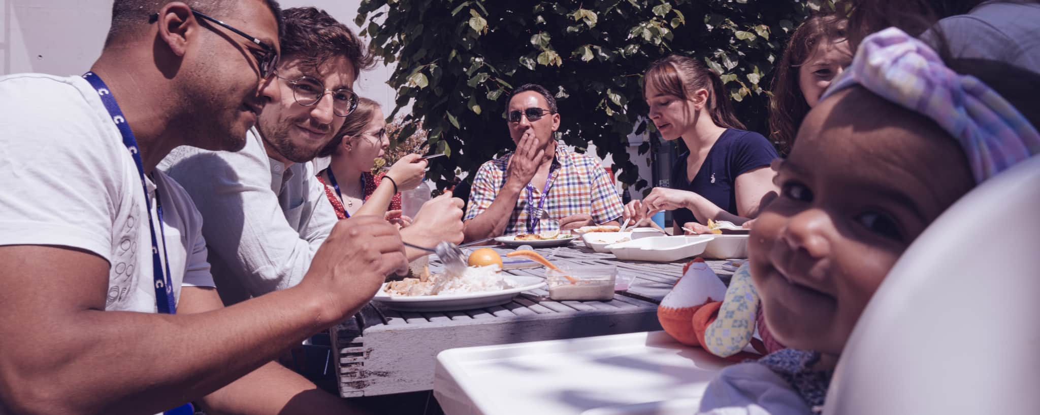 Several people sat around a table enjoying lunch, a baby at the table is looking over its shoulder at the camera.