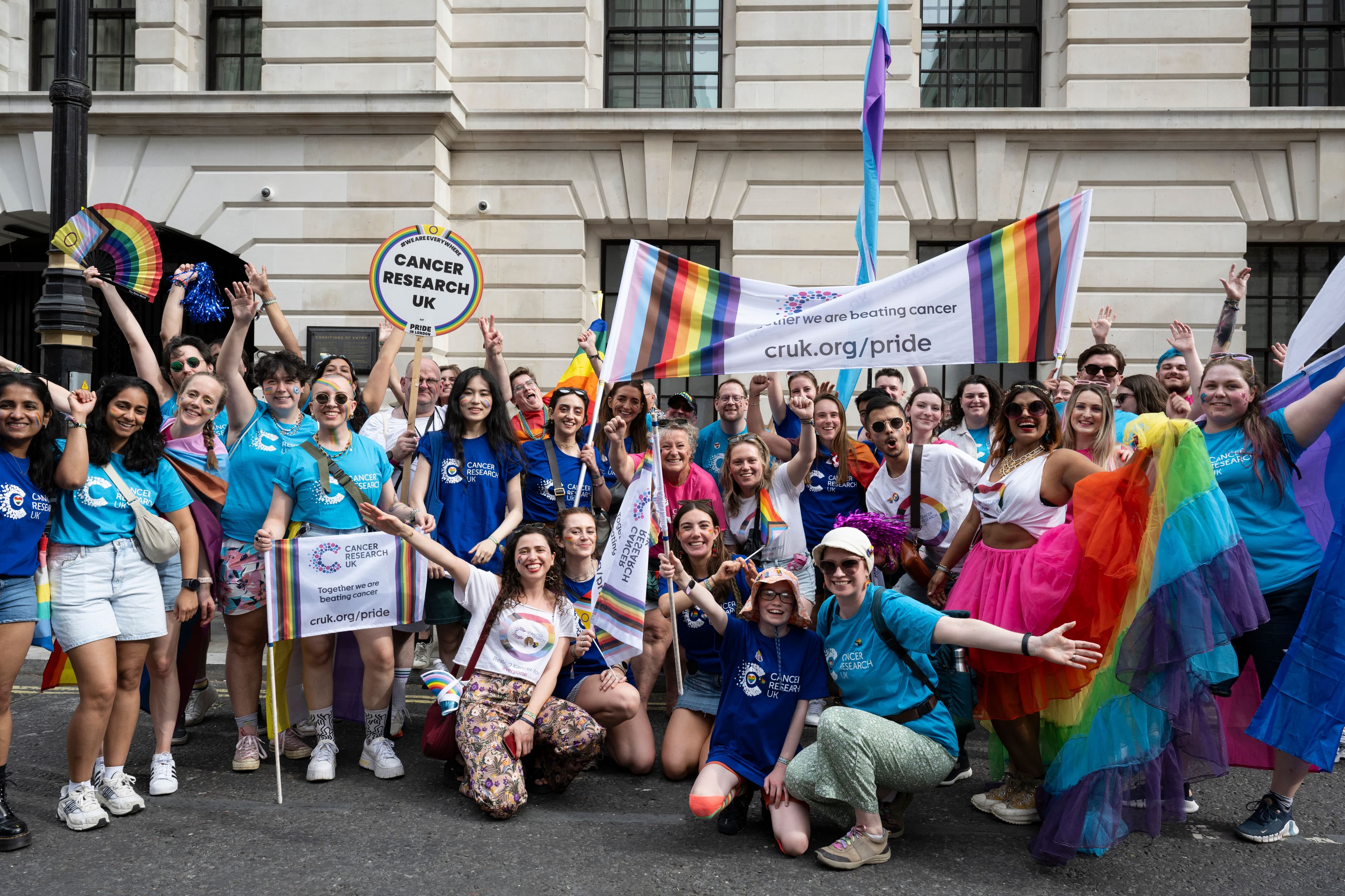 A large group of people wearing cancer research UK t-shirts, cheering and waving LGBTQIA flags.
