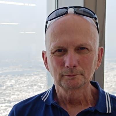 A person with sunglasses on top of their head, wearing a blue polo shirt, standing in front of a window in a tall building.