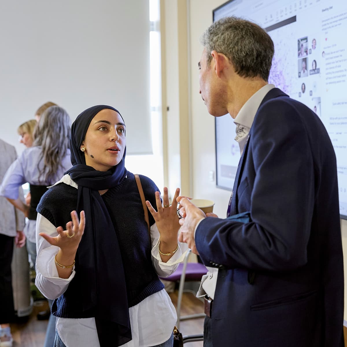 Two people having a discussion at an event. They are standing in front of a screen.