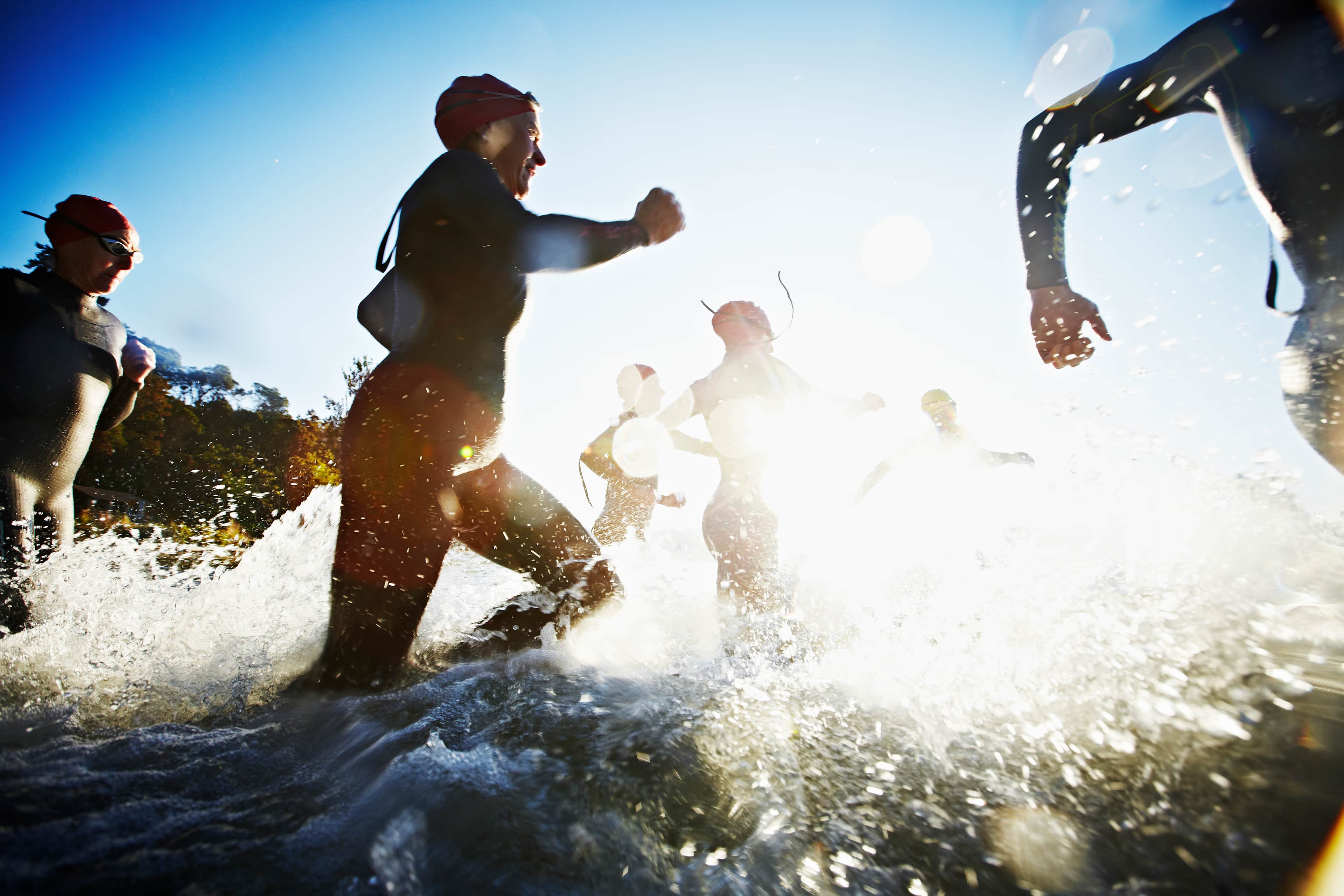 Group of mature triathletes wearing wetsuits swim caps and goggles running into water at sunrise.