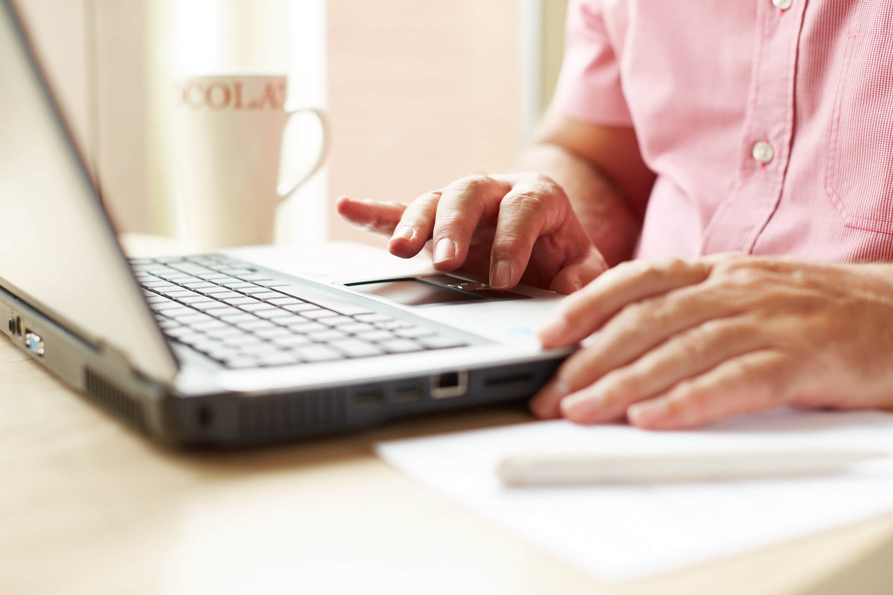 A man is sat at a table, using a laptop. There is a cup of tea besides the laptop.