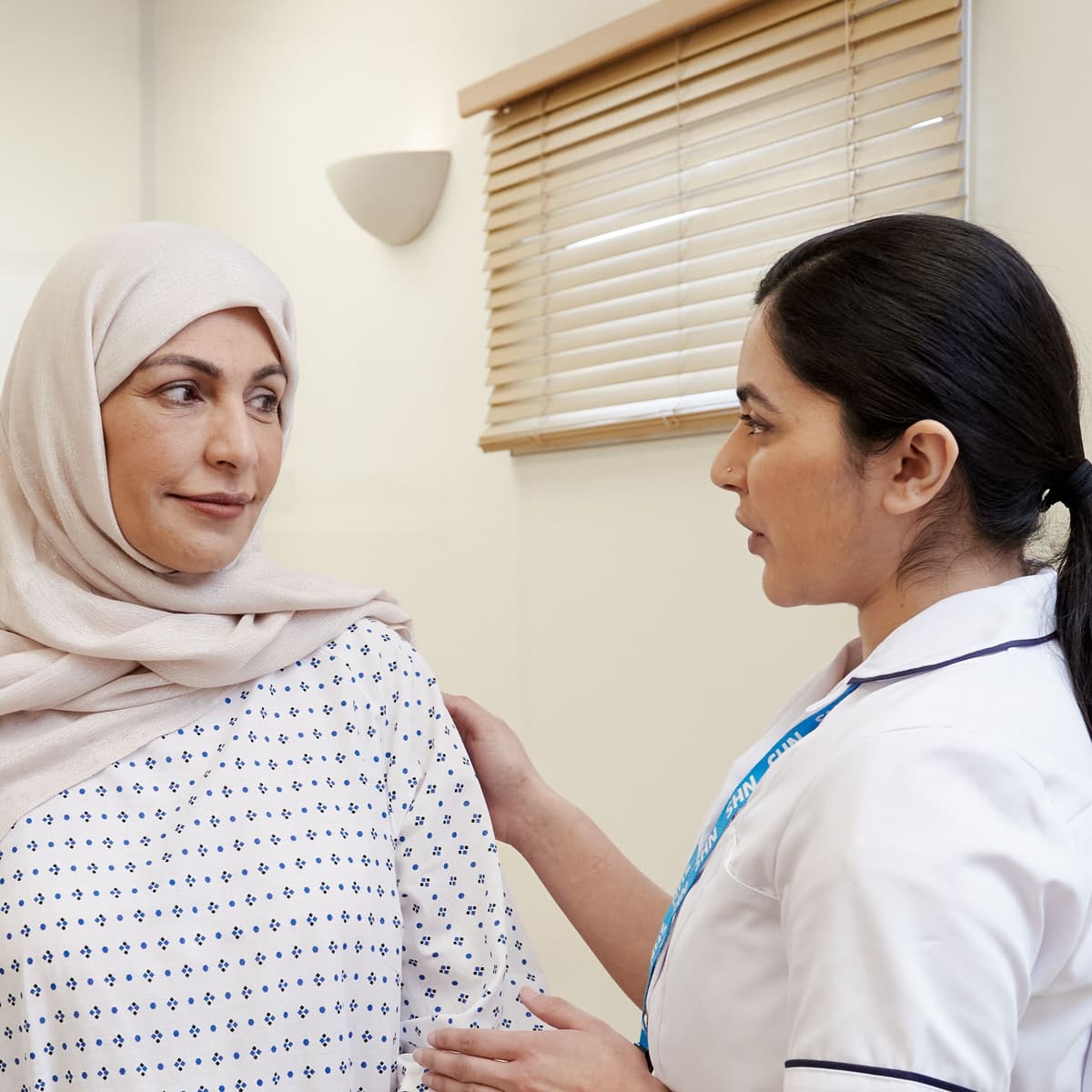 This photo shows a mammographer with a patient, wearing a hospital gown, before a mammogram.