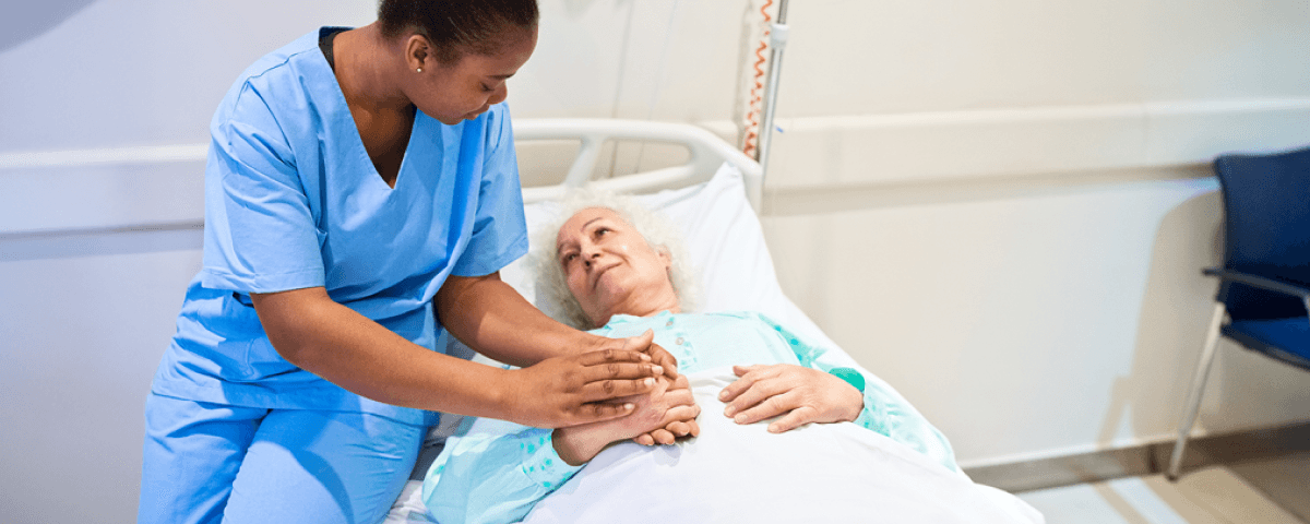 A female doctor sitting on a female patient's bed in a hospital ward, holding her hand.