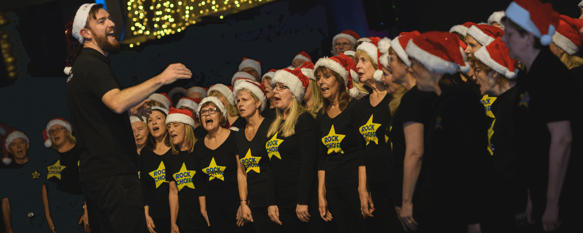 Choir singing with festive hats in front of supporters at a Legacy Festive Music event.