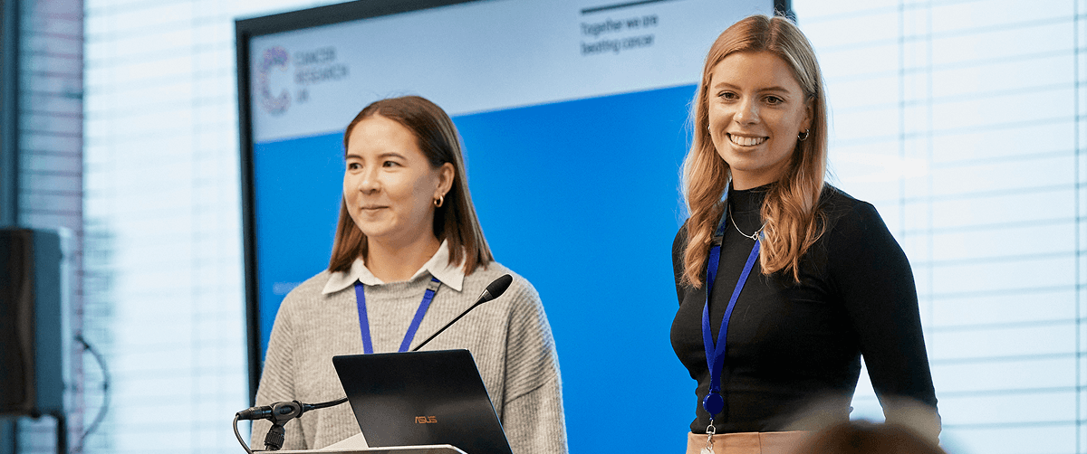 Two people standing in front of a video screen giving a presentation.