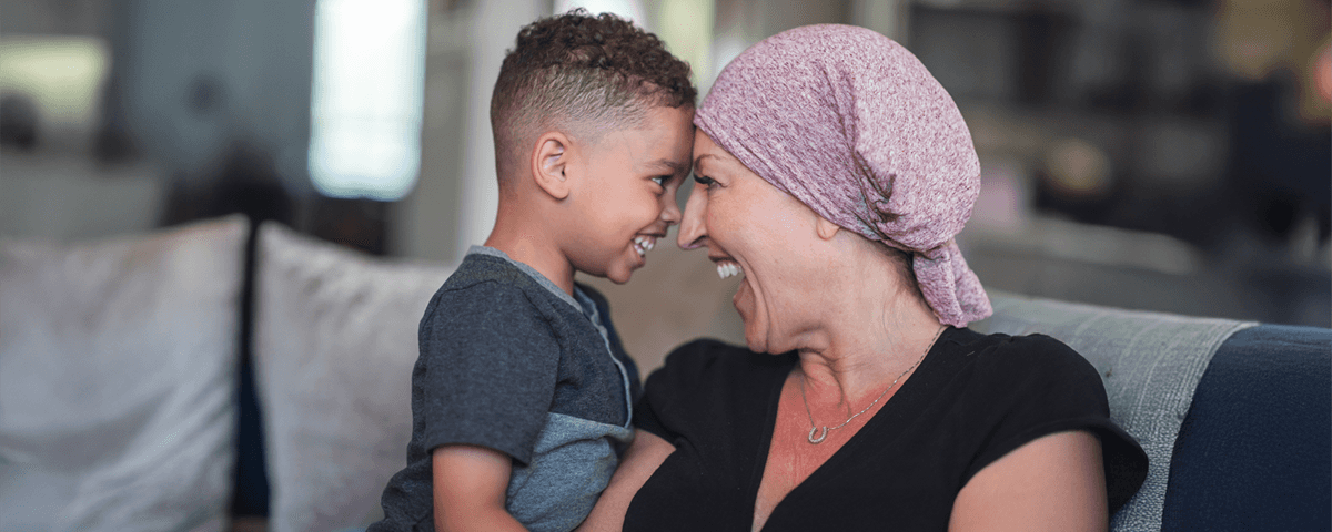 A woman wearing a head wrap is holding a small boy. Their foreheads are touching and they are smiling broadly at each other.