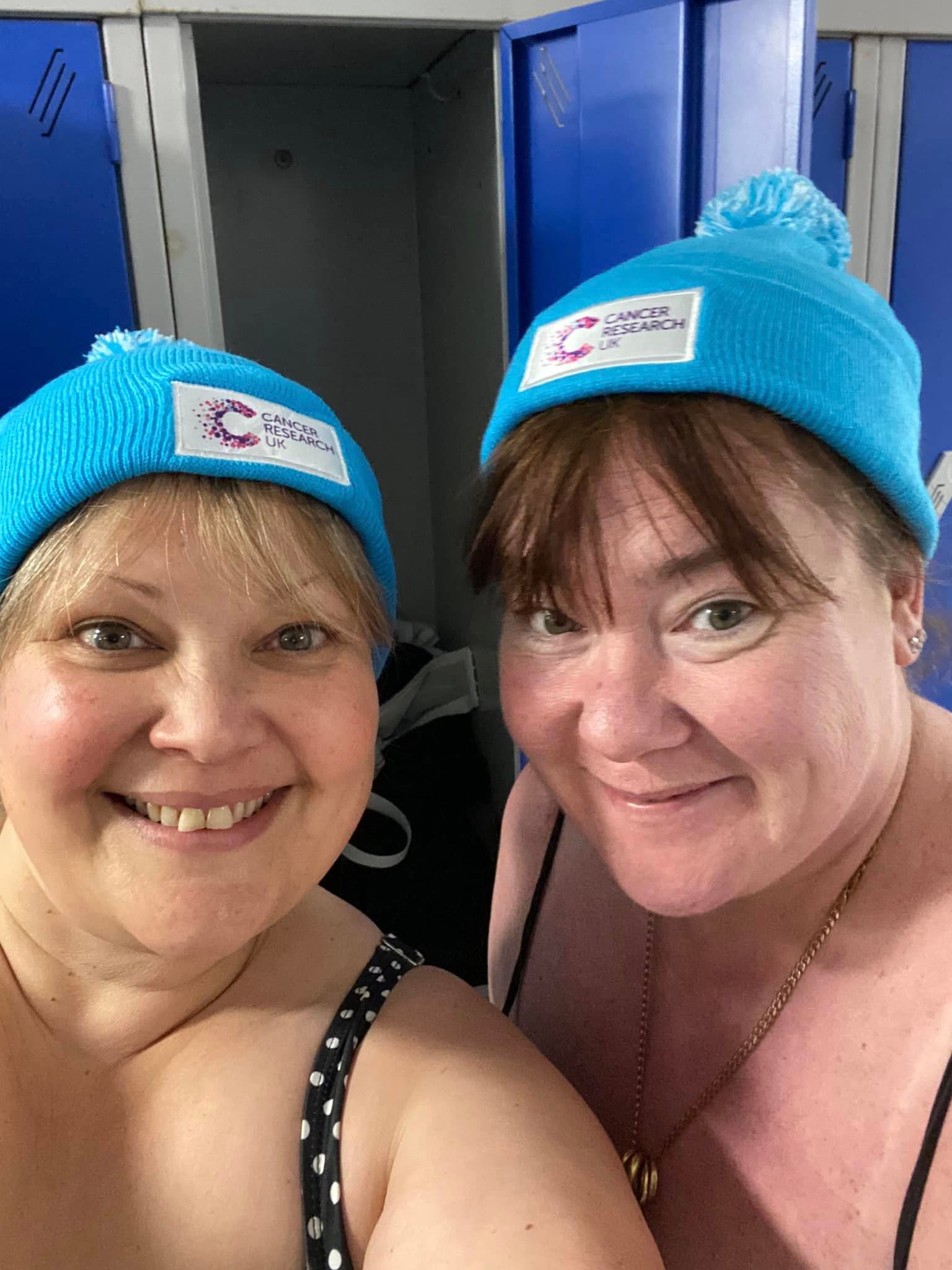 Two women smiling in a swimming pool changing room wearing the Cancer Research UK bobble hat.
