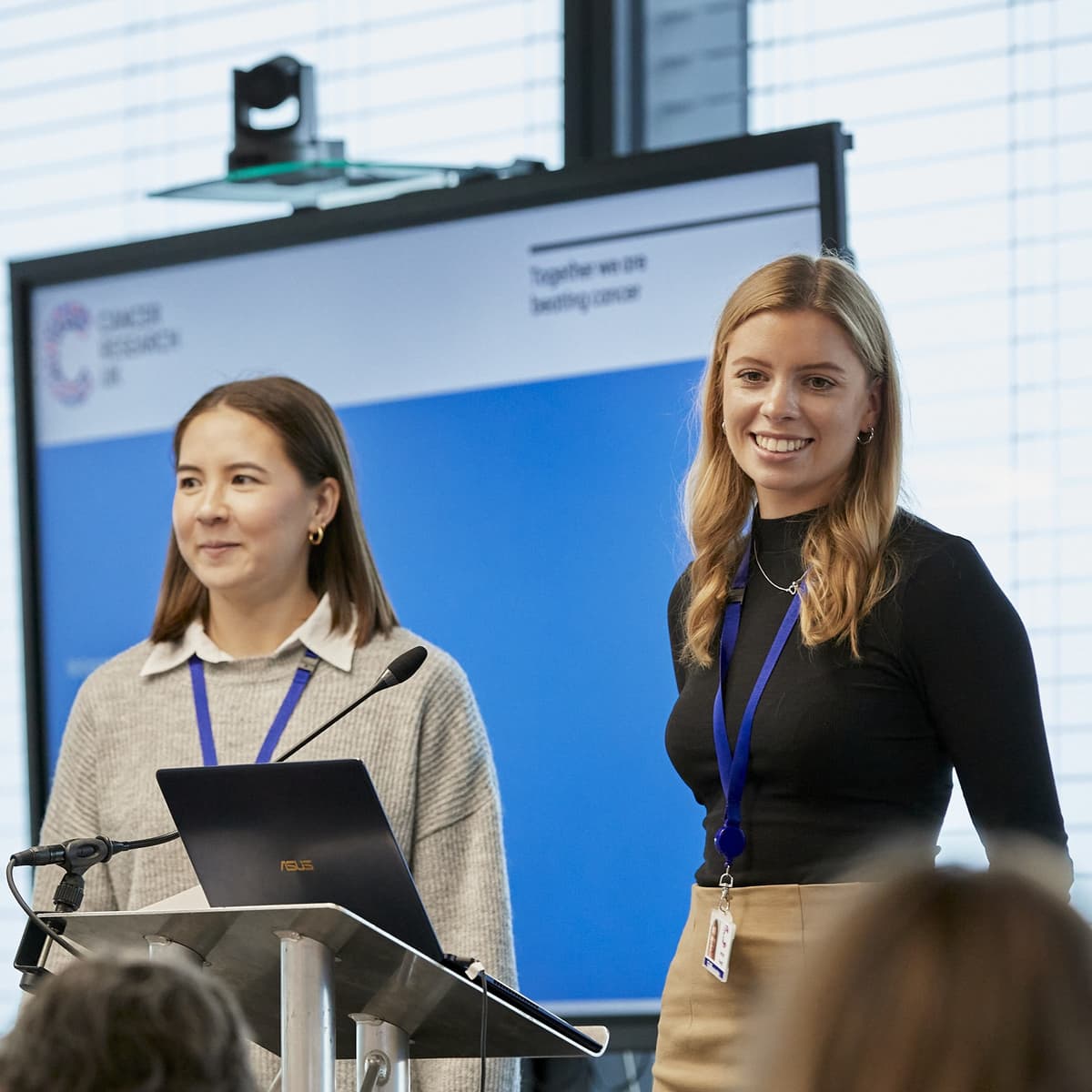 Two people standing in front of a video screen giving a presentation.