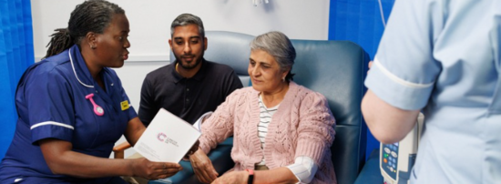 Patient is sat with a family member and a health professional while having chemo. A nurse shows them a Cancer Research UK leaflet.