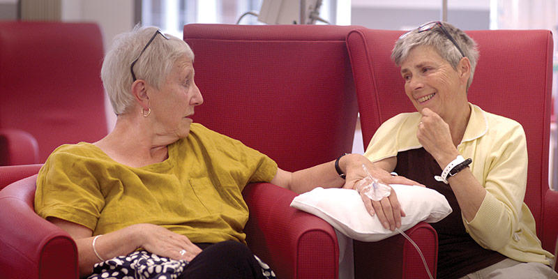 Two women talk and laugh while one receives cancer treatment