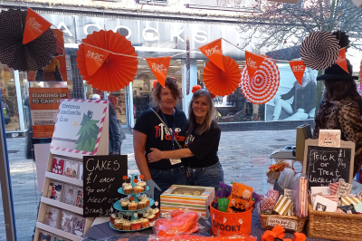 Two women in front of a Stand Up to Cancer stall set up in a shop