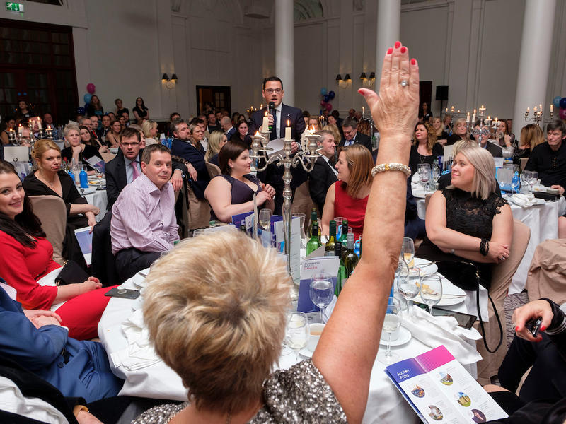 a room full of people at an event sitting at their tables doing an auction, with a woman raising her hand