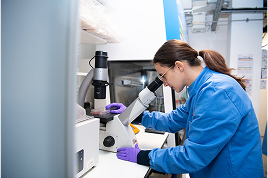 image of a female researcher wearing a blue lab coat and looking into a microscope