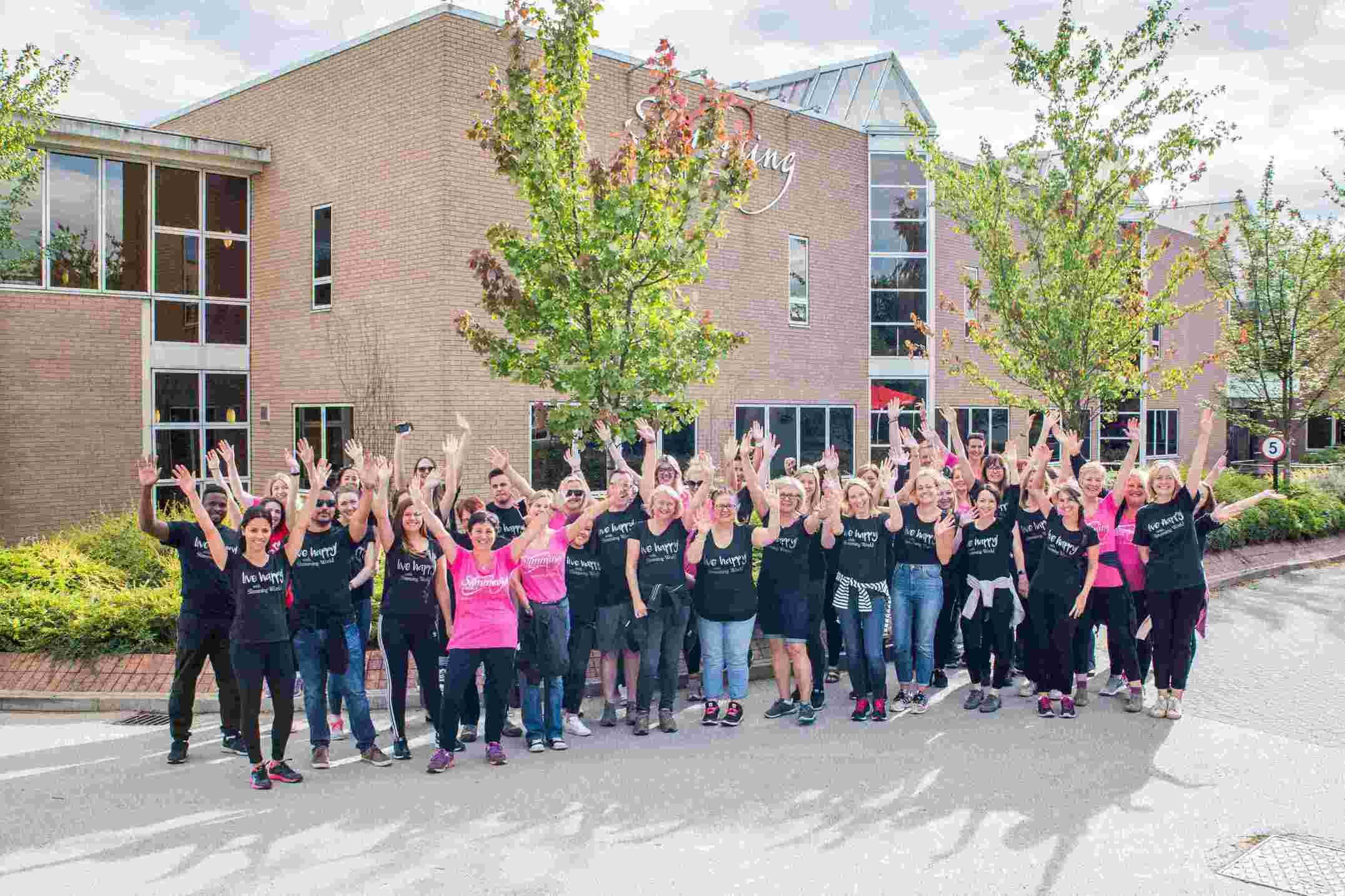 Group of people wearing pink or black Slimming World T-Shirts with their hands in the air and smiling in front of a Slimming World building