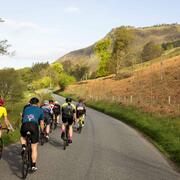 A group of cyclists (photographed from behind), cycling a country road with a backdrop of the Scottish highlands